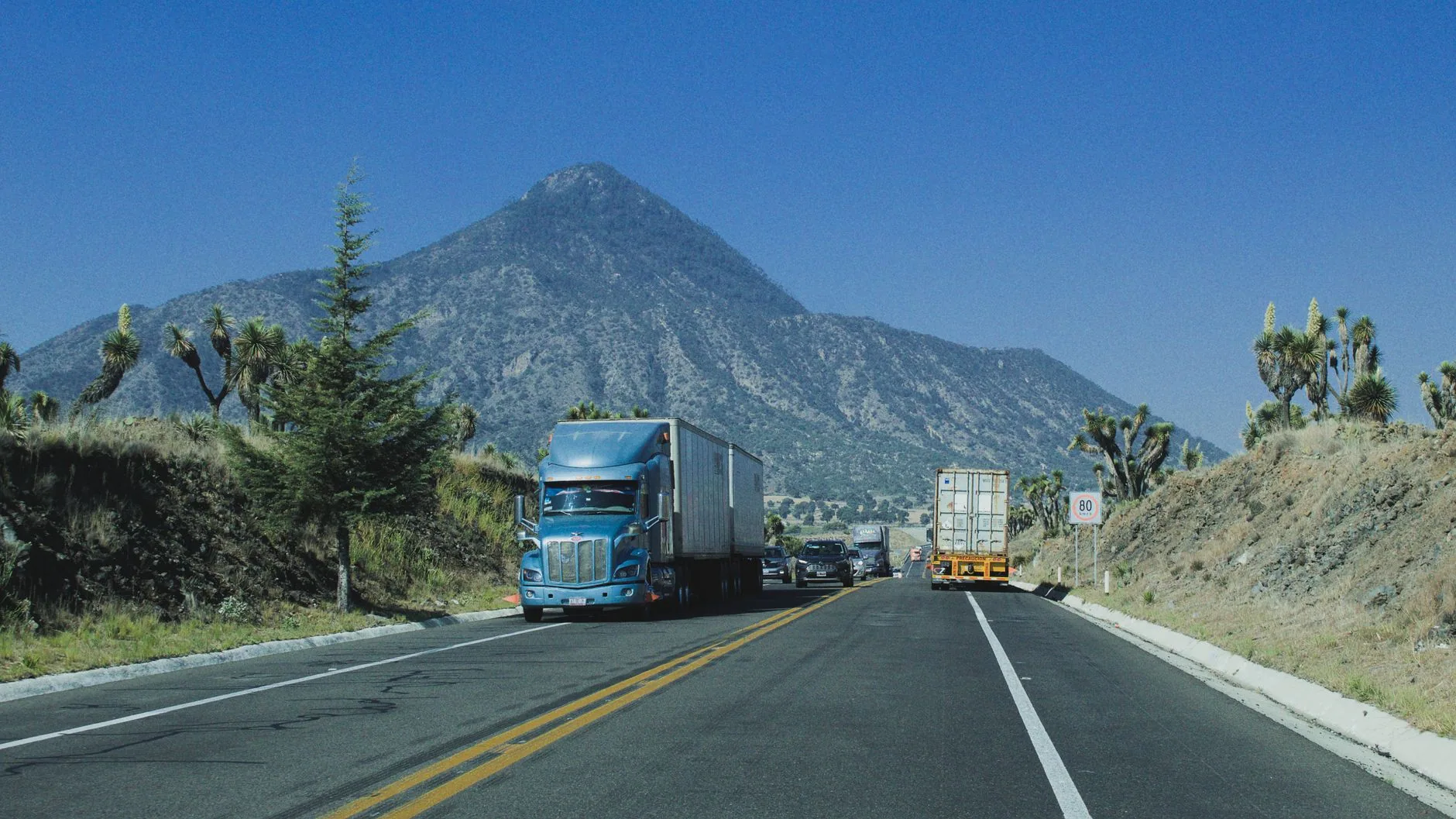 Tractocamión azul con caja seca circula por una carretera de dos carriles frente a un cerro