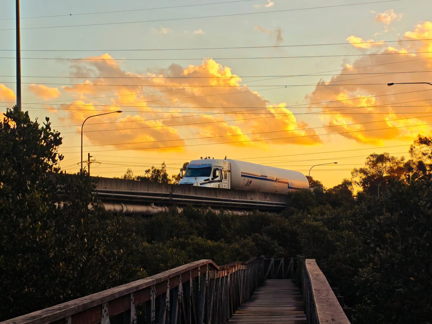 Pipa de combustible cruza un puente carretero al atardecer, entre árboles y cables de luz