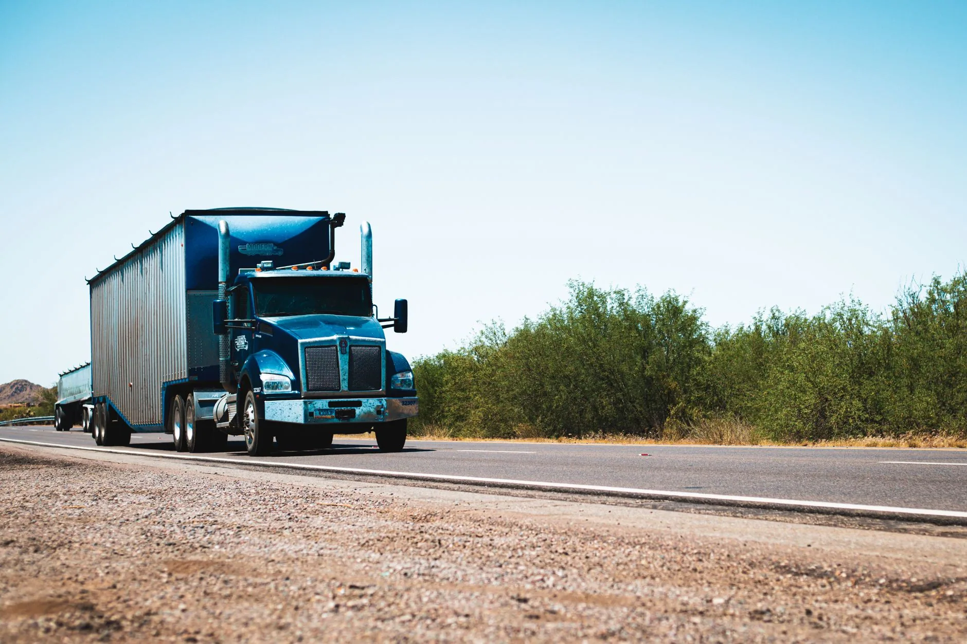 Tractocamión azul con remolque circulando por una carretera rodeada de matorrales