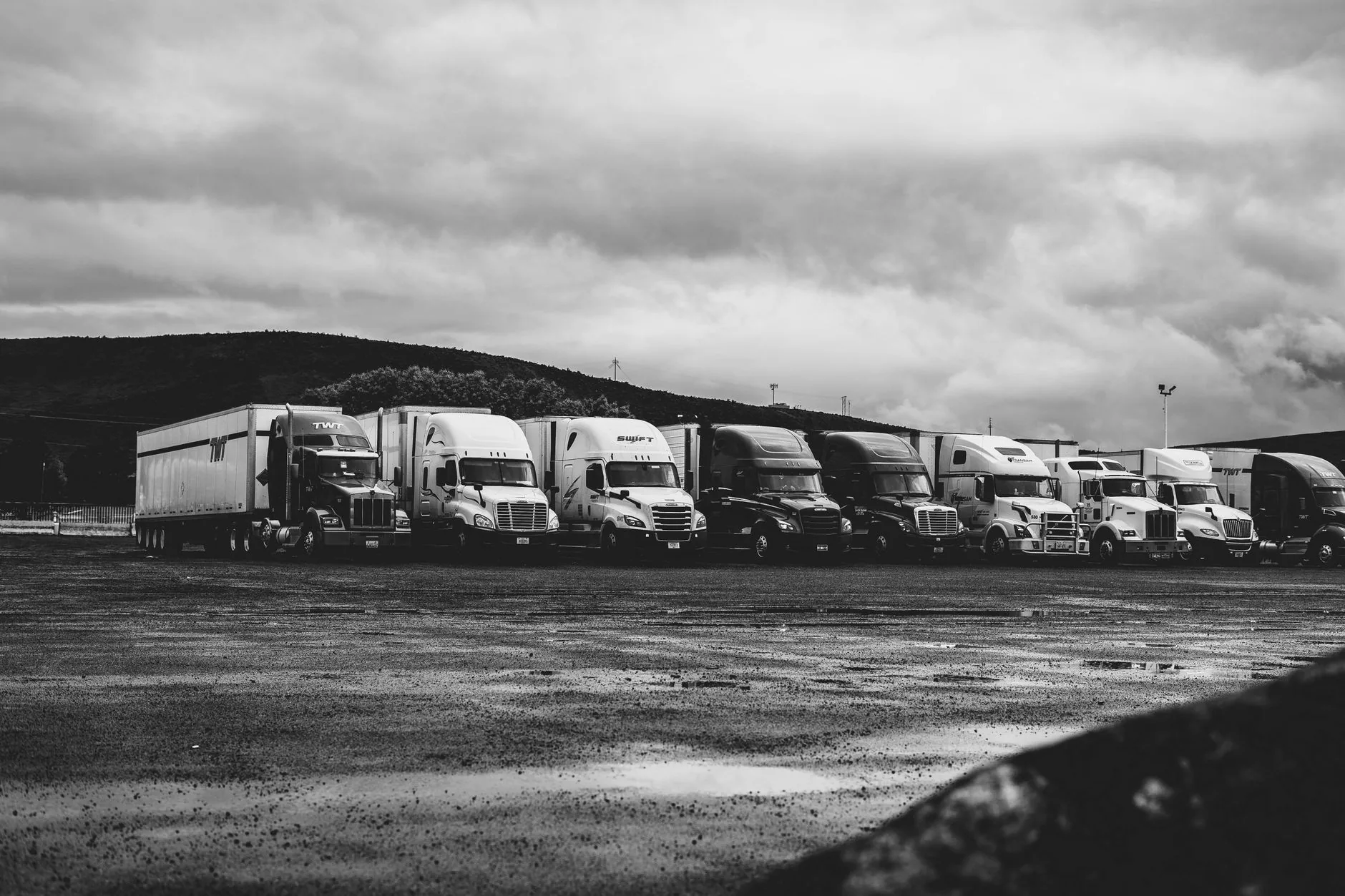Fila de tractocamiones estacionados en un patio de terracería bajo un cielo nublado, en blanco y negro