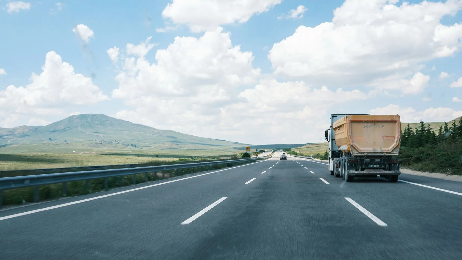 Camión de volteo con caja naranja circulando por una autopista de cuatro carriles con cerros al fondo