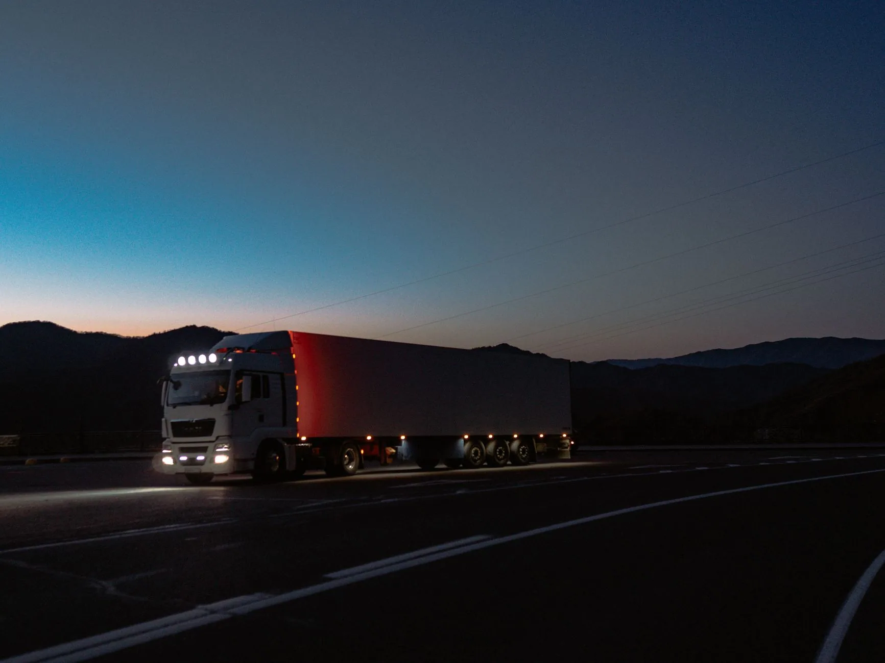 Tractocamión con caja seca y luces encendidas en una carretera al anochecer, con cerros al fondo